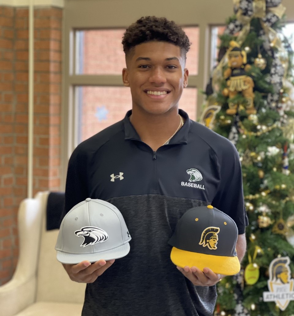 Raebert De Los Santos stands while holding a Central Methodist baseball hat in one hand and a FTCC baseball hat in the other hand.