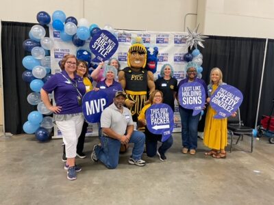 A group of volunteers and Theo the FTCC Mascot pose for a photo in front of a United Way backdrop.