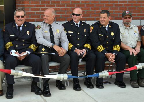 A group of fire and emergency response dignitaries sit behind a fire hose.