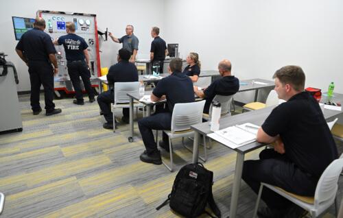 Students sit looking at four men standing at an instrument panel in a simulation lab.